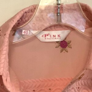 May include: A pale pink blouse with floral embroidery and eyelet detailing. The blouse has a pointed collar and is on a clear plastic hanger. The label reads "PINK BOUTIQUE" with a floral design. Size 40.