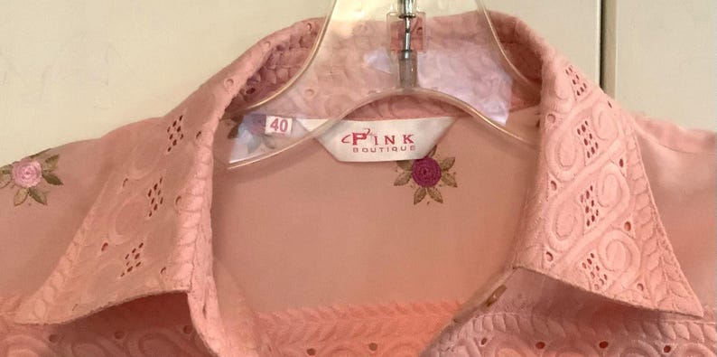 May include: A pale pink blouse with floral embroidery and eyelet detailing. The blouse has a pointed collar and is on a clear plastic hanger. The label reads "PINK BOUTIQUE" with a floral design. Size 40.