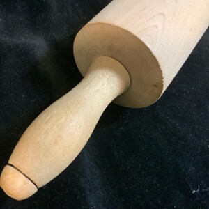 May include: A light-colored wooden rolling pin with a cylindrical body and a handle. The handle is attached to the body with a rounded joint. The rolling pin is made of a light-colored wood, and the grain of the wood is visible.