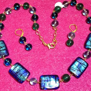 May include: A blue and green glass necklace with matching earrings. The necklace features rectangular glass beads with iridescent blue and green colors. The necklace is strung on a gold chain.