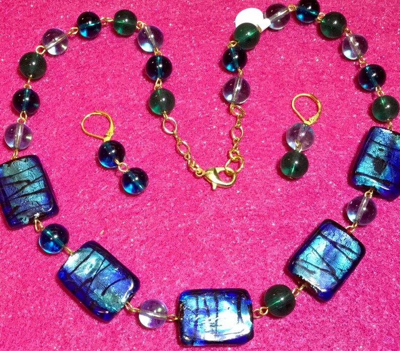May include: A blue and green glass necklace with matching earrings. The necklace features rectangular glass beads with iridescent blue and green colors. The necklace is strung on a gold chain.