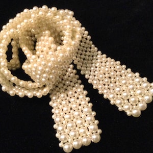May include: A long, flexible belt or sash constructed from numerous small, cream-colored faux pearls woven together in a dense, grid-like pattern. The accessory features a continuous, interconnected structure of spherical beads with a smooth finish.