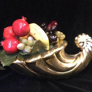 May include: A gold cornucopia fruit bowl filled with artificial fruit, including red pomegranates, green grapes, a yellow banana, and dark red cherries.