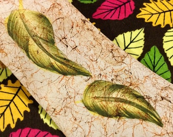 May include: A beige tie with a textured surface and two green leaves printed on it. The tie is shown against a brown background with colorful leaves.