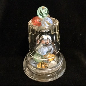 May include: A clear glass jar with a wide mouth, filled with colorful marbles. The marbles are a mix of blue, green, red, and white. The jar is sitting on a clear glass base.