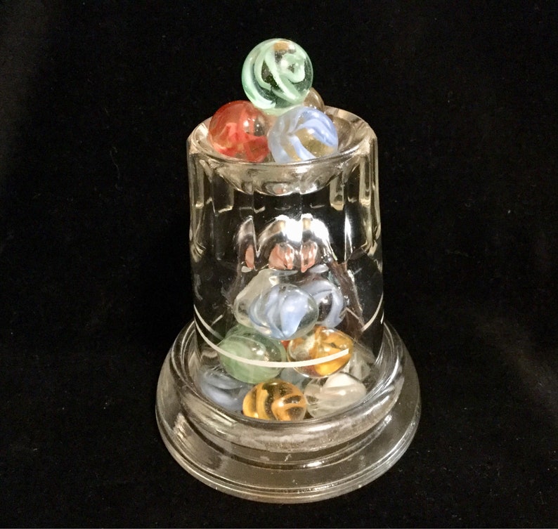 May include: A clear glass jar with a wide mouth, filled with colorful marbles. The marbles are a mix of blue, green, red, and white. The jar is sitting on a clear glass base.
