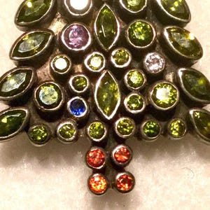 May include: A silver Christmas tree charm with green, red, blue, and purple rhinestones. The charm is decorated with a variety of small, colorful stones.