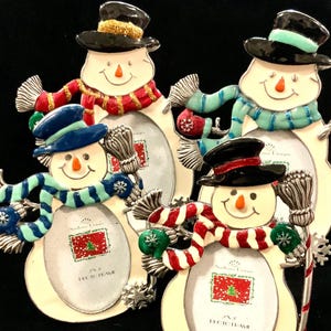 May include: Four snowman-shaped picture frames with a white background and a silver metal frame. Each snowman has a different colored scarf and hat. The frames are decorated with a candy cane, a snowflake, and a broom.