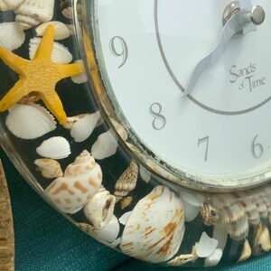 May include: A decorative clock with a white face and silver hands, featuring the text "Sands of Time." The clock's clear frame is filled with seashells and a yellow starfish, creating a coastal theme.