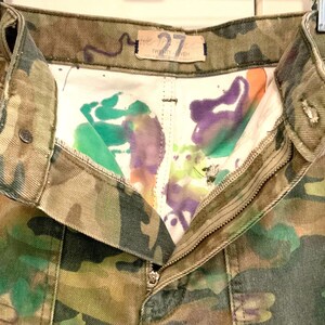 May include: Close-up of camouflage-patterned pants with a partially unzipped zipper. The interior lining features colorful paint splatters in green, purple, and orange. The pants have a button closure and a small tag that reads "27".