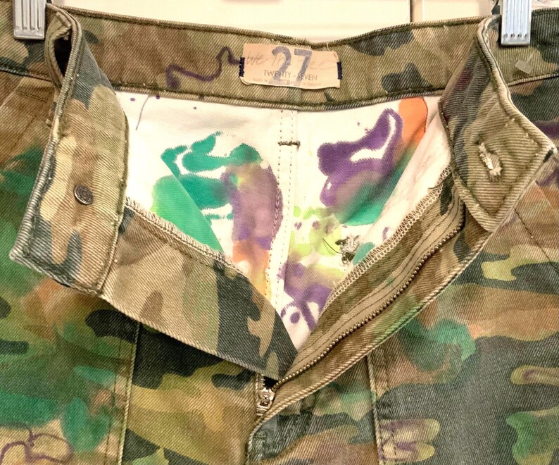May include: Close-up of camouflage-patterned pants with a partially unzipped zipper. The interior lining features colorful paint splatters in green, purple, and orange. The pants have a button closure and a small tag that reads "27".