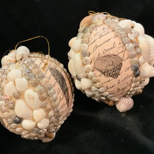 May include: Two white Christmas ornaments covered in seashells and small white beads. The ornaments have a vintage, handwritten design with the text "Stick Around" and "Free Dollars".