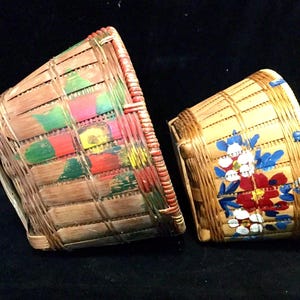 May include: Two woven baskets with painted floral designs. The larger basket has green, red, yellow, and white flowers. The smaller basket has blue, red, white, and yellow flowers.