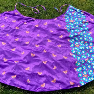 Op de afbeelding: Een paarse stoffen cape met een vlinderprint. De cape heeft aan de ene kant een bloemenprint en aan de andere kant een effen paarse kleur.