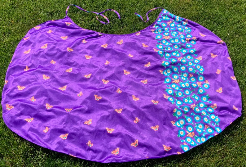 Op de afbeelding: Een paarse stoffen cape met een vlinderprint. De cape heeft aan de ene kant een bloemenprint en aan de andere kant een effen paarse kleur.
