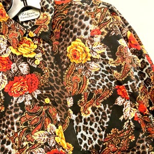 May include: Button-down shirt with a vibrant floral and paisley pattern. The shirt features a mix of red, yellow, and orange roses, set against a black background with leopard print accents. The label reads "California Connection Inc."