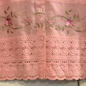 May include: A pale pink blouse with floral embroidery and eyelet detailing. The blouse has a button-down front and short sleeves. The embroidery features pink roses and green leaves. The bottom hem has a scalloped edge.