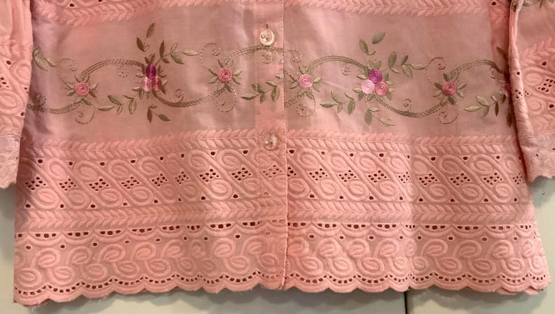May include: A pale pink blouse with floral embroidery and eyelet detailing. The blouse has a button-down front and short sleeves. The embroidery features pink roses and green leaves. The bottom hem has a scalloped edge.