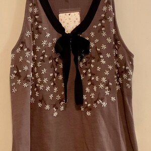 May include: A brown sleeveless top with a black bow and white floral embroidery. The top has a black trim around the neckline.