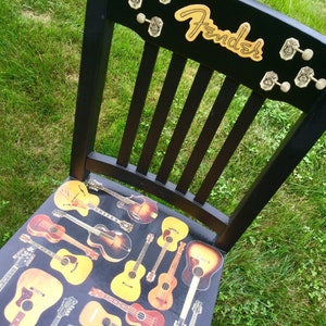 Op de afbeelding: Een zwarte houten stoel met een Fender-logo op de rugleuning. De zitting is bedekt met een collage van vintage gitaren.