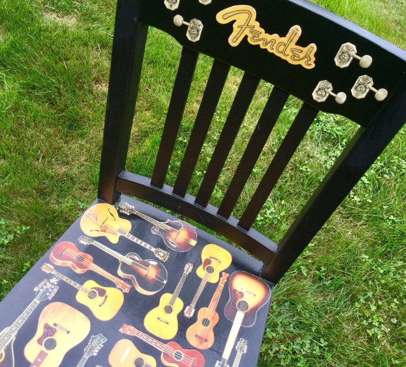 Op de afbeelding: Een zwarte houten stoel met een Fender-logo op de rugleuning. De zitting is bedekt met een collage van vintage gitaren.