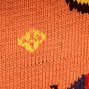 K&ouml;nnte beinhalten: Nahaufnahme eines gestrickten Stoffes mit einem bunten geometrischen Muster. Der Stoff ist haupts&auml;chlich orange mit gelben, schwarzen, blauen und roten Akzenten.
