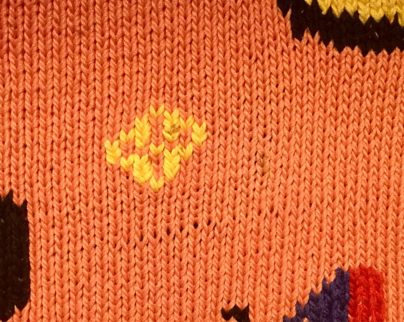 K&ouml;nnte beinhalten: Nahaufnahme eines gestrickten Stoffes mit einem bunten geometrischen Muster. Der Stoff ist haupts&auml;chlich orange mit gelben, schwarzen, blauen und roten Akzenten.