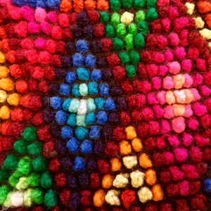 K&ouml;nnte beinhalten: Nahaufnahme eines bunten gewebten Teppichs mit geometrischem Muster. Der Teppich besteht aus kleinen, runden, flauschigen Garnkugeln in den Farben Rot, Rosa, Orange, Gelb, Gr&uuml;n, Blau und Lila.