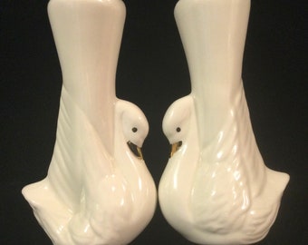 May include: A pair of white ceramic dove-shaped vases with gold accents. The vases are facing each other and have a tall, cylindrical opening.