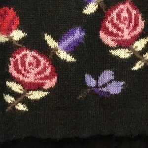 May include: Black knitted sweater with a floral pattern featuring pink, red, and purple flowers. The flowers are embroidered on the sweater.