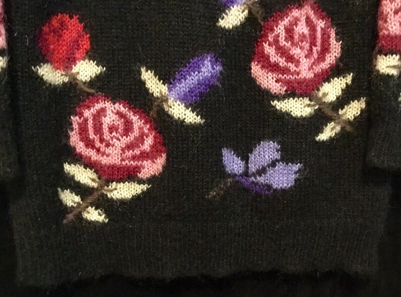May include: Black knitted sweater with a floral pattern featuring pink, red, and purple flowers. The flowers are embroidered on the sweater.