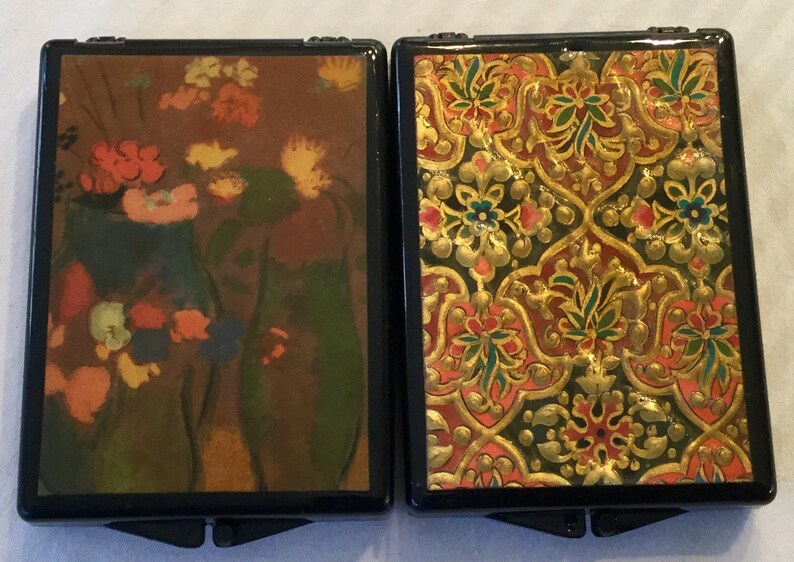 May include: Two small, rectangular boxes with black frames. One features a floral still life in shades of orange, yellow, and blue. The other has a gold, red, and green ornate pattern.