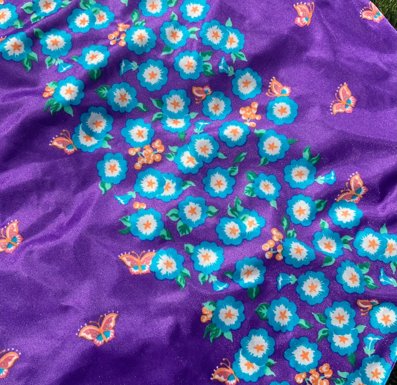 Op de afbeelding: Paarse stof met een herhalend patroon van blauwe bloemen met witte centra en groene bladeren. Er zijn ook oranje vlinders verspreid over de stof.