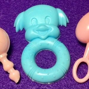 Pode incluir: Tr&ecirc;s brinquedos para beb&eacute;s: um chocalho rosa, um anel azul com uma cara sorridente e um mordedor em forma de cora&ccedil;&atilde;o rosa.