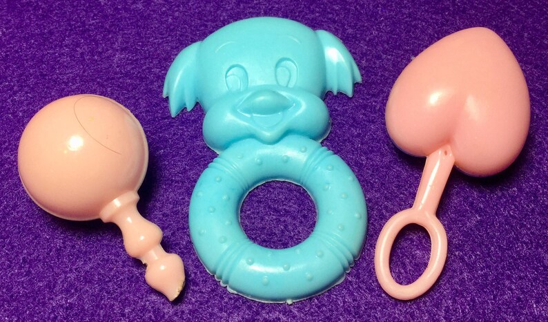Pode incluir: Tr&ecirc;s brinquedos para beb&eacute;s: um chocalho rosa, um anel azul com uma cara sorridente e um mordedor em forma de cora&ccedil;&atilde;o rosa.