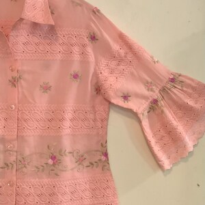 May include: A pale pink shirt with eyelet and floral embroidery. The shirt has three-quarter length sleeves with a flared design and floral accents. The collar is visible, and the fabric appears lightweight and breathable.