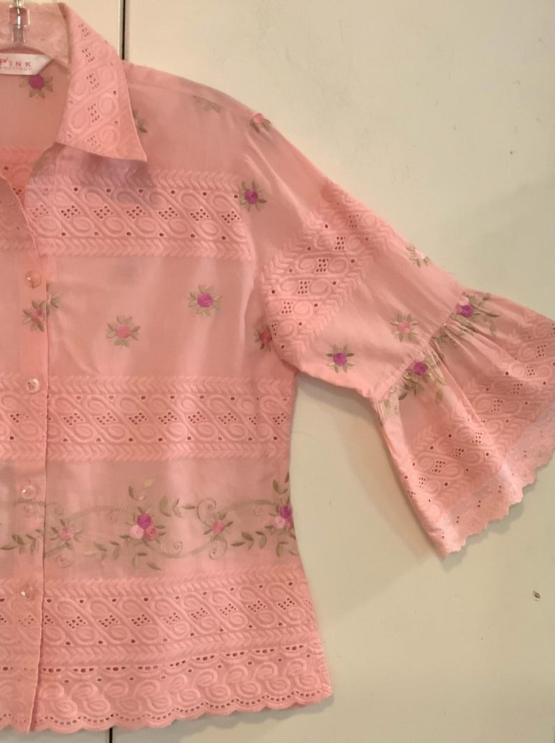 May include: A pale pink shirt with eyelet and floral embroidery. The shirt has three-quarter length sleeves with a flared design and floral accents. The collar is visible, and the fabric appears lightweight and breathable.