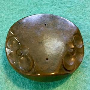 May include: A round, dark brown, ceramic incense burner with a raised design of three circles on each side. The burner has two small holes on the top.