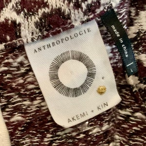 Puede incluir: Una etiqueta blanca con texto negro que dice "Anthropologie" y "Akemi + Kin". La etiqueta tambi&eacute;n presenta un c&iacute;rculo negro con l&iacute;neas radiantes. La etiqueta est&aacute; unida a una tela estampada de color burdeos y blanco con purpurina plateada.
