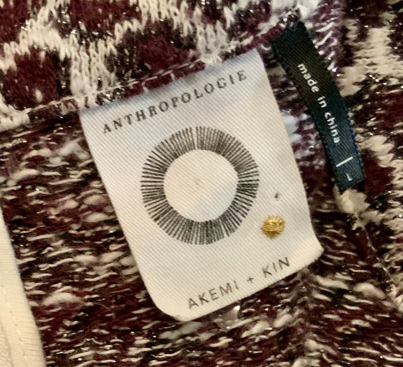 Puede incluir: Una etiqueta blanca con texto negro que dice "Anthropologie" y "Akemi + Kin". La etiqueta tambi&eacute;n presenta un c&iacute;rculo negro con l&iacute;neas radiantes. La etiqueta est&aacute; unida a una tela estampada de color burdeos y blanco con purpurina plateada.