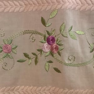 May include: Pale pink sheer fabric with embroidered floral and leaf design. The embroidery features clusters of pink and purple roses with green leaves and stems. The top and bottom edges have a decorative leaf pattern.