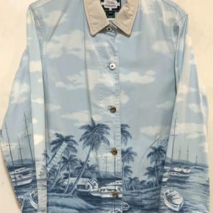 May include: Light blue button-up jacket with a beige collar. The jacket features a scenic print of palm trees, boats, and a coastal landscape. The sky is depicted with white clouds. The jacket has silver buttons.