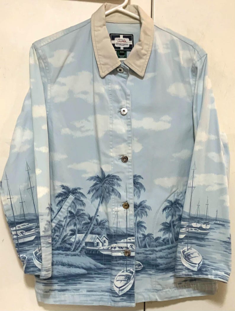 May include: Light blue button-up jacket with a beige collar. The jacket features a scenic print of palm trees, boats, and a coastal landscape. The sky is depicted with white clouds. The jacket has silver buttons.