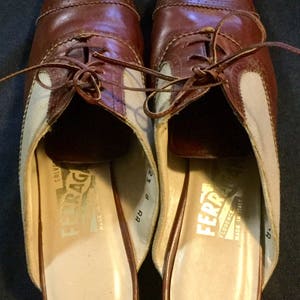 May include: A pair of brown leather and beige canvas slip-on shoes with laces. The shoes have a brown leather sole and are labelled "Salvatore Ferragamo Florence Italy Made in Italy" on the inside.