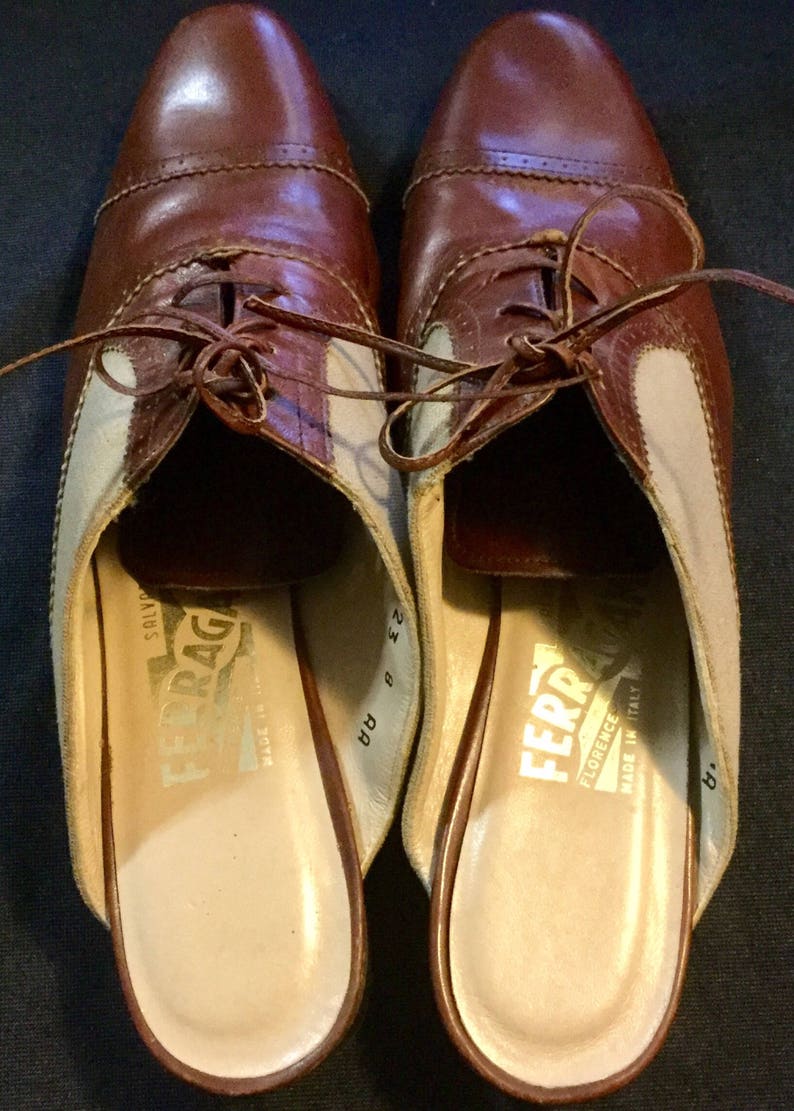 May include: A pair of brown leather and beige canvas slip-on shoes with laces. The shoes have a brown leather sole and are labelled "Salvatore Ferragamo Florence Italy Made in Italy" on the inside.
