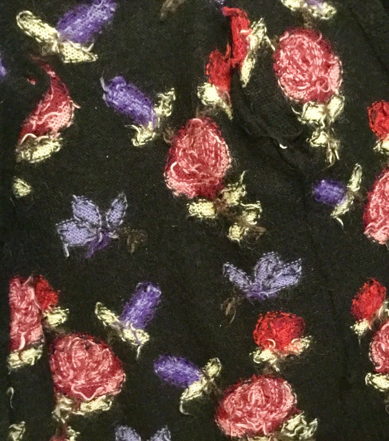 May include: Black fabric with a repeating pattern of pink, red, and purple flowers. The flowers are embroidered and have a fuzzy texture.