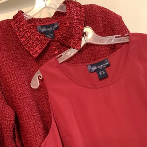 May include: Two red tops with a textured and smooth fabric. The tops have a label that reads "Susan Gravely Jenkins" and a size XL.