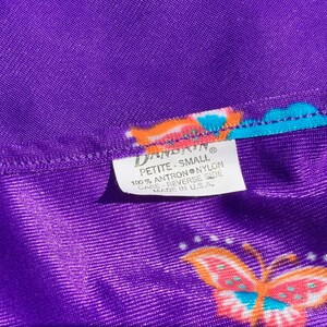 Op de afbeelding: Paarse stof met een vlinderprint en een wit label met de tekst "DANDAIN PETITE-SMALL 100% ANTRON NYLON CARE REVERSE SIDE MADE IN U.S.A."