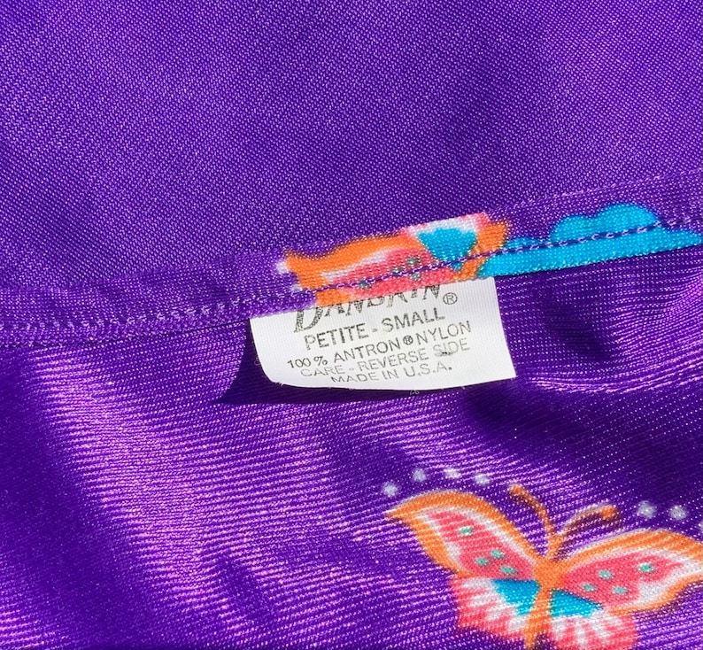 Op de afbeelding: Paarse stof met een vlinderprint en een wit label met de tekst "DANDAIN PETITE-SMALL 100% ANTRON NYLON CARE REVERSE SIDE MADE IN U.S.A."