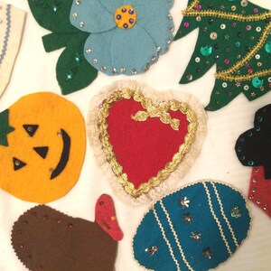 May include: A collection of felt ornaments in various shapes and colors, including a pumpkin, a heart, a Christmas tree, a flower, an egg, and a turkey. The ornaments are decorated with sequins, glitter, and other embellishments.